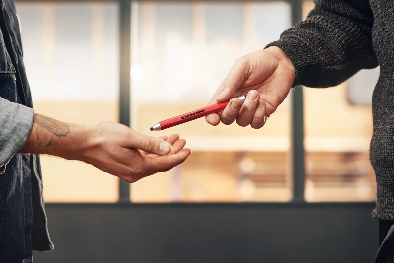 Es sind Hände von zwei Personen zu sehen welche sich einen Edding-Stift überreichen.