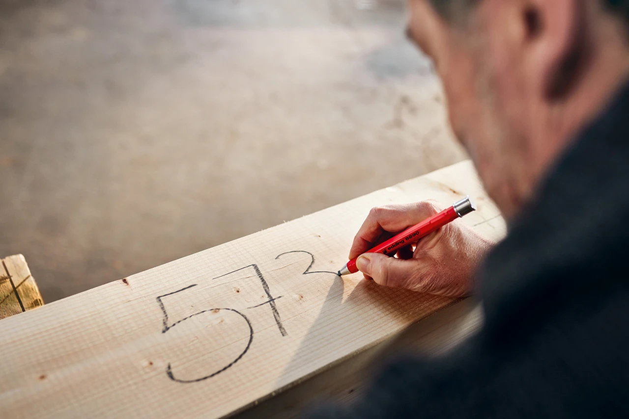 Ein Mann wird in einem Over-the-Shoulder-Shot fotografiert, wie er mit einem edding eine Zahl auf einen Holzbalken schreibt