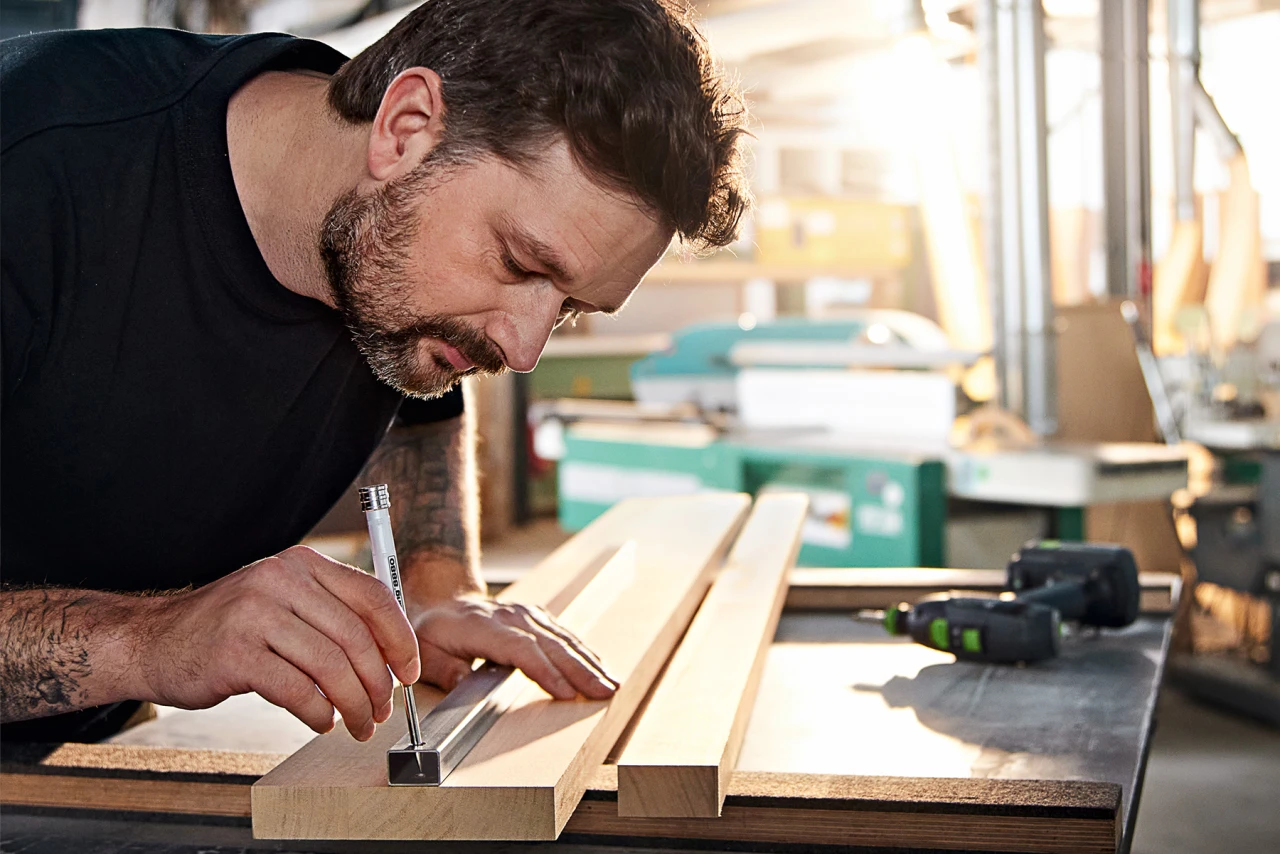 Ein Mann wird in einer Nahaufnahme fotografiert, wie er mithilfe eines eddings eine Bohrmarkierung durch ein Loch in einem Metallbalken anzeichnet